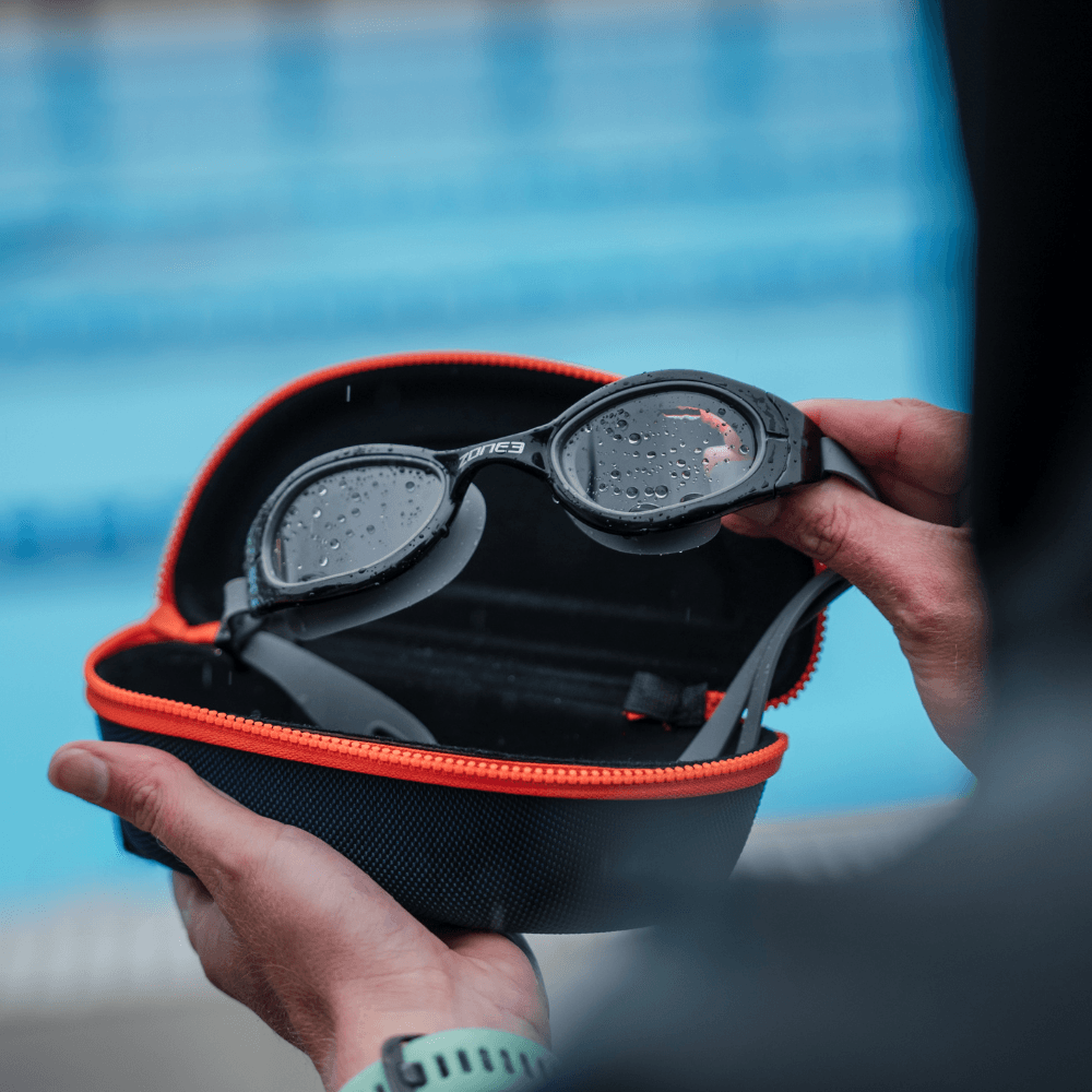 Estuche de gafas azul marino abierto, mostrando unas gafas de natación Zone3 con gotas de agua en las lentes.