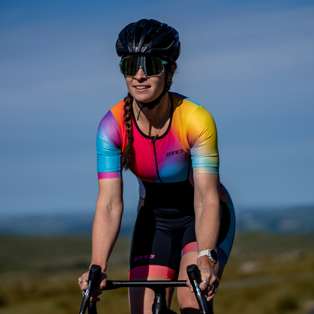 Primer plano frontal de una ciclista con el trisuit Activate+ de manga corta para mujer de ZONE3. Los colores degradados en azul, rosa, amarillo y naranja son visibles bajo la luz del sol.