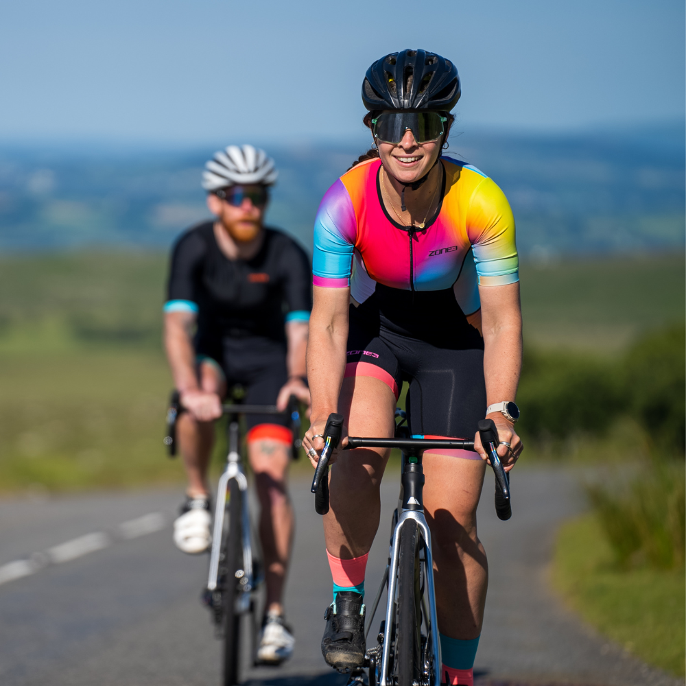 Mujer pedaleando al aire libre con el trisuit Activate+ de manga corta para mujer de ZONE3, casco negro y gafas de sol. El diseño degradado es visible en hombros y brazos.