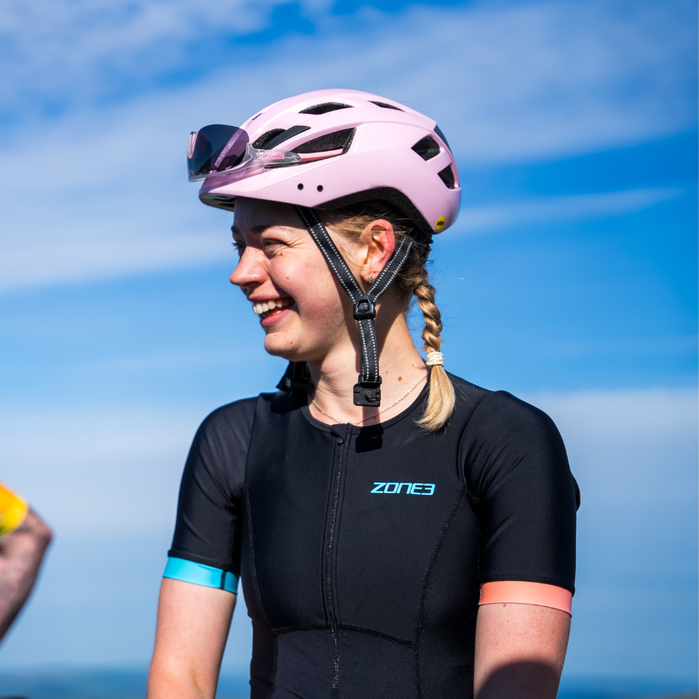 Mujer con el trisuit Activate+ de manga corta para mujer de ZONE3 en negro, con puños de manga en rosa y azul. Sonríe y lleva un casco rosa.