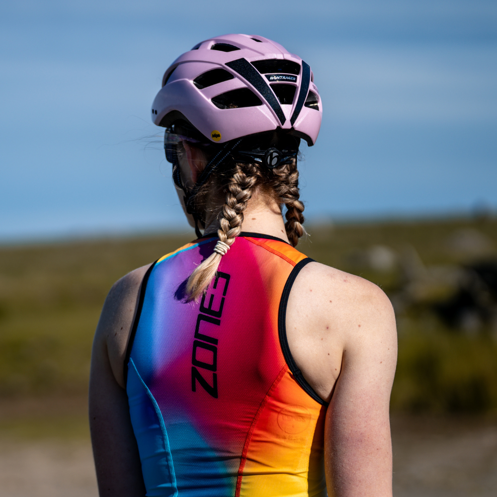 Ciclista con el trisuit Activate+ sin mangas de ZONE3 y casco rosa. El trisuit tiene el logotipo vertical de ZONE3 y un vivo diseño degradado en la espalda.